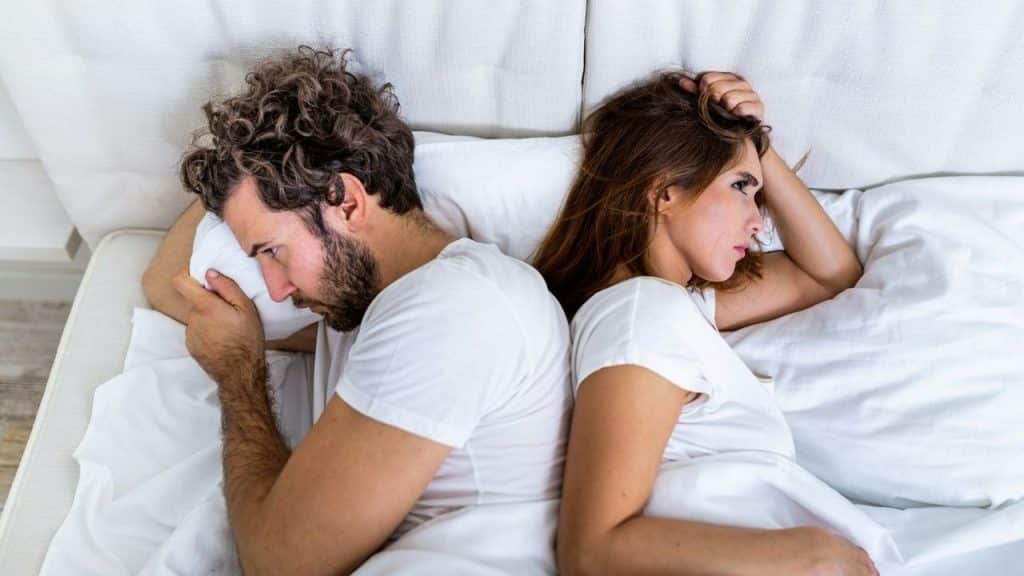 A man and woman sitting back-to-back on a bed looking away with frustrated expressions.