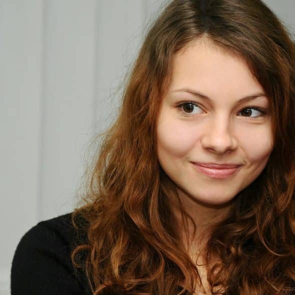 A close-up portrait of a smiling young woman with wavy brown hair looking to the side indoors.