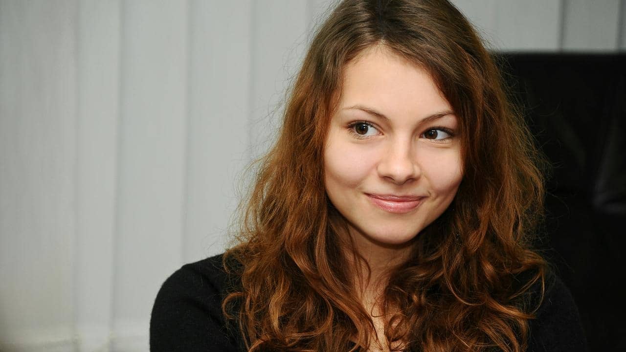 A close-up portrait of a smiling young woman with wavy brown hair looking to the side indoors.