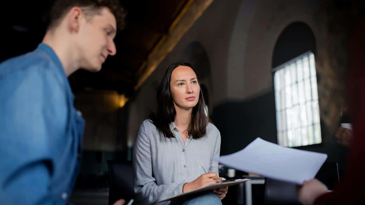 A woman holds a clipboard while listening during a meeting with coworkers indoors.
