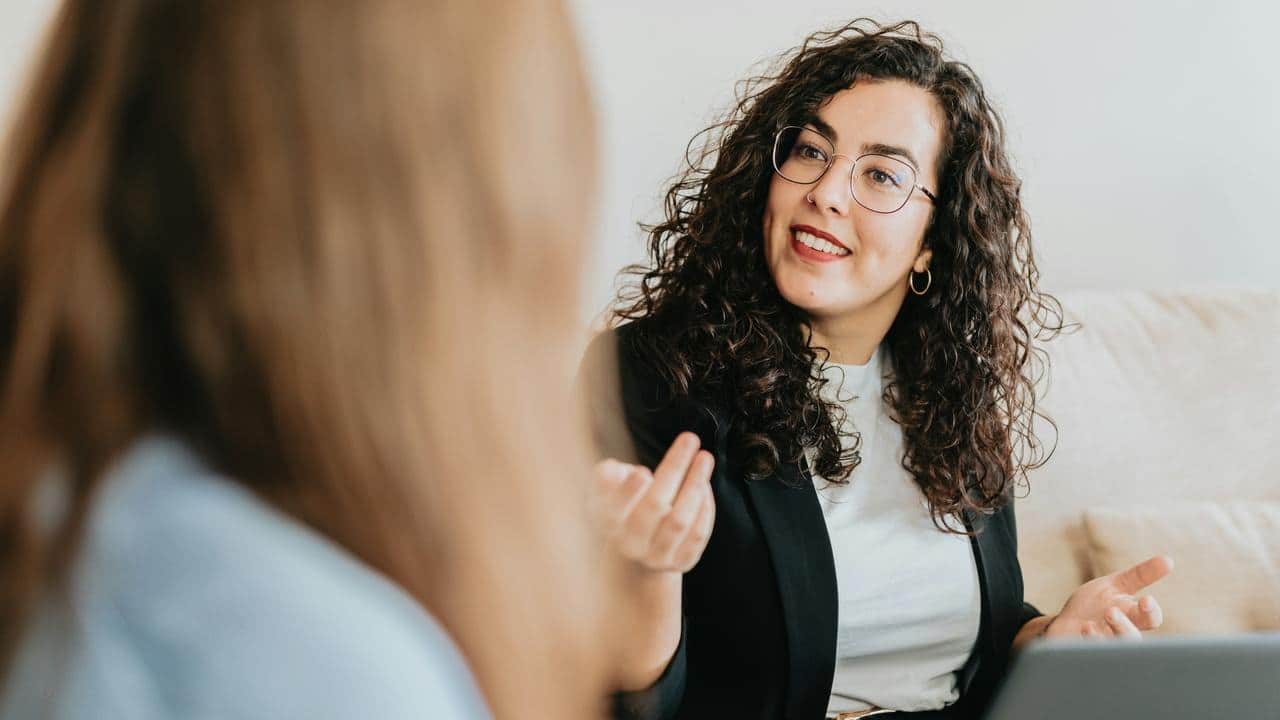 A smiling woman with curly hair and glasses talks while gesturing during a conversation.