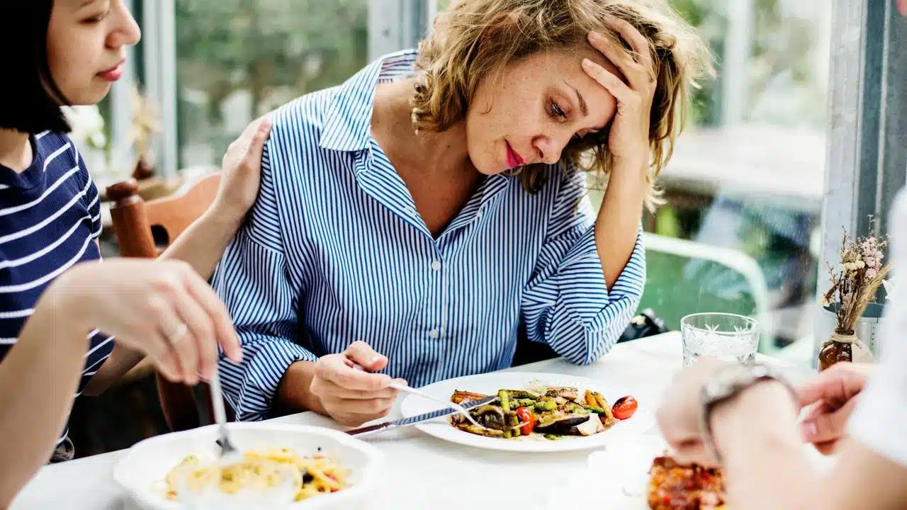 A distressed woman sits at a table with food while another person comforts her.