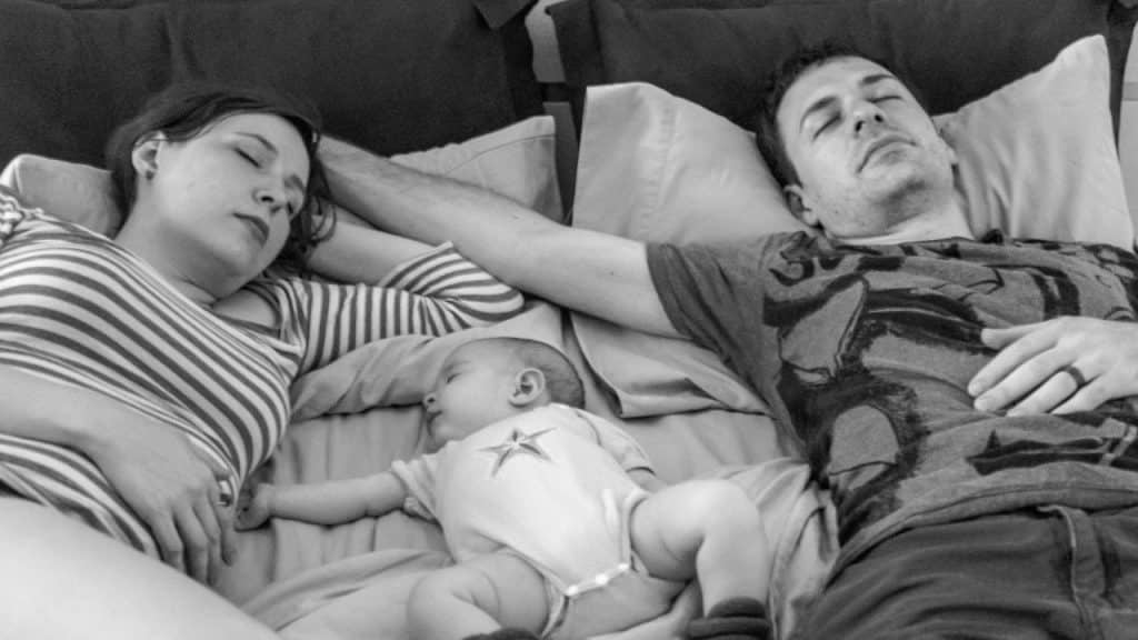 A black and white overhead photo of two adults and an infant sleeping on a bed.