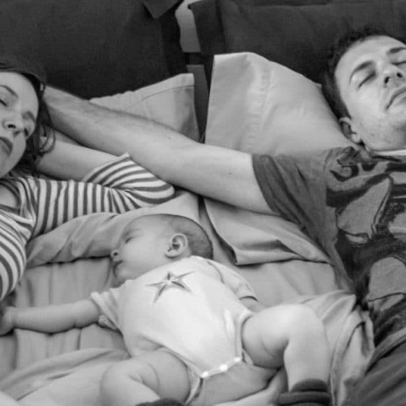 A black and white overhead photo of two adults and an infant sleeping on a bed.