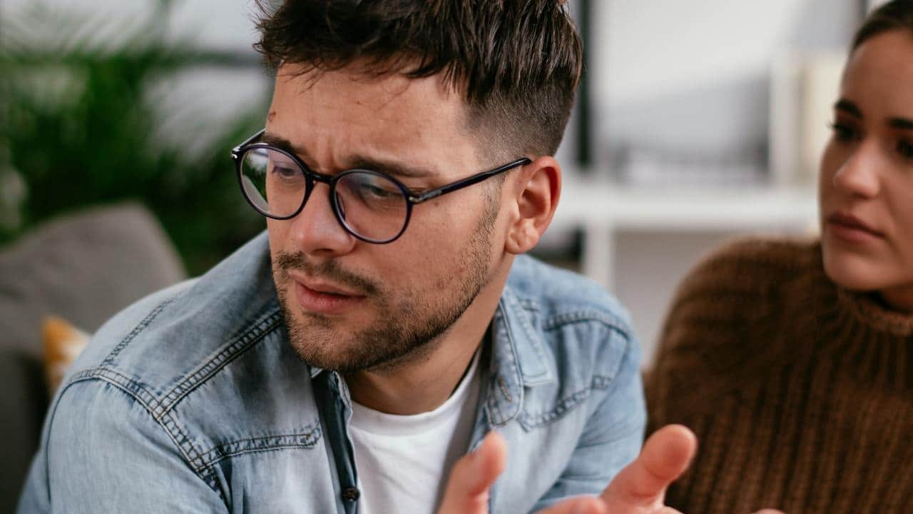 A worried man wearing glasses talks while a woman listens in the background.