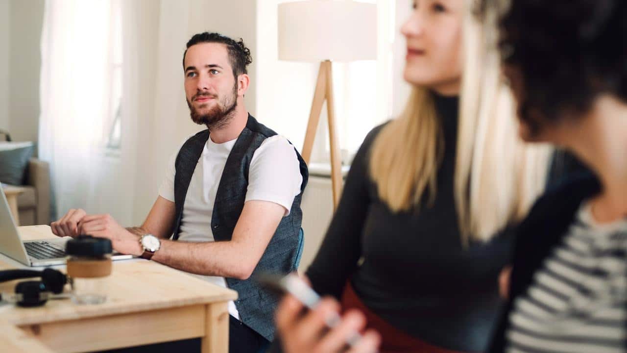 A man sits at a desk with a laptop while two people talk nearby.