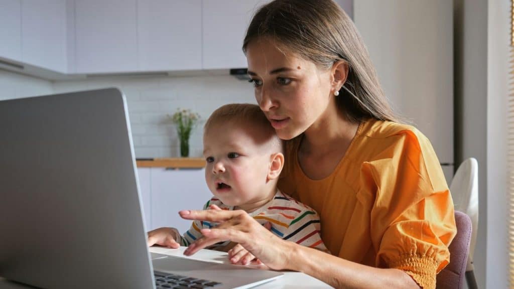 A woman in an orange shirt sits at a laptop with a young child on her lap.
