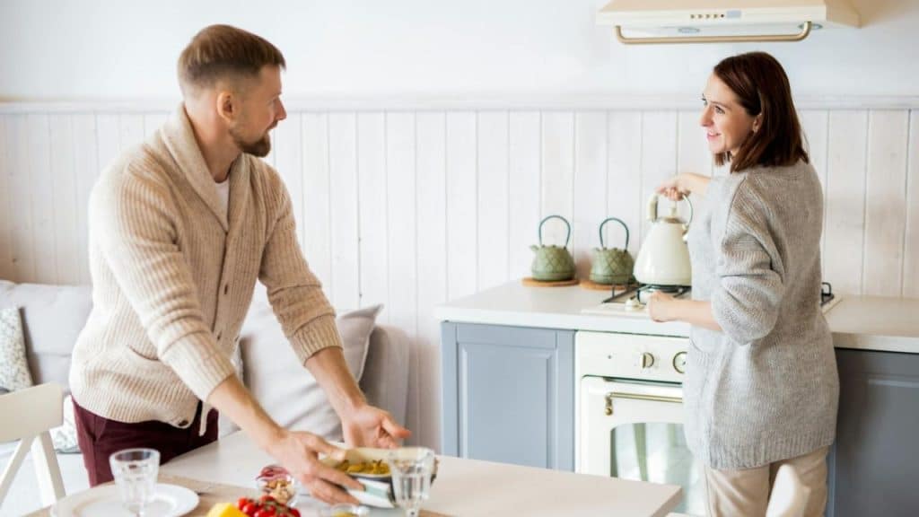 A man places a bowl on a table while a woman stands by a kitchen stove.