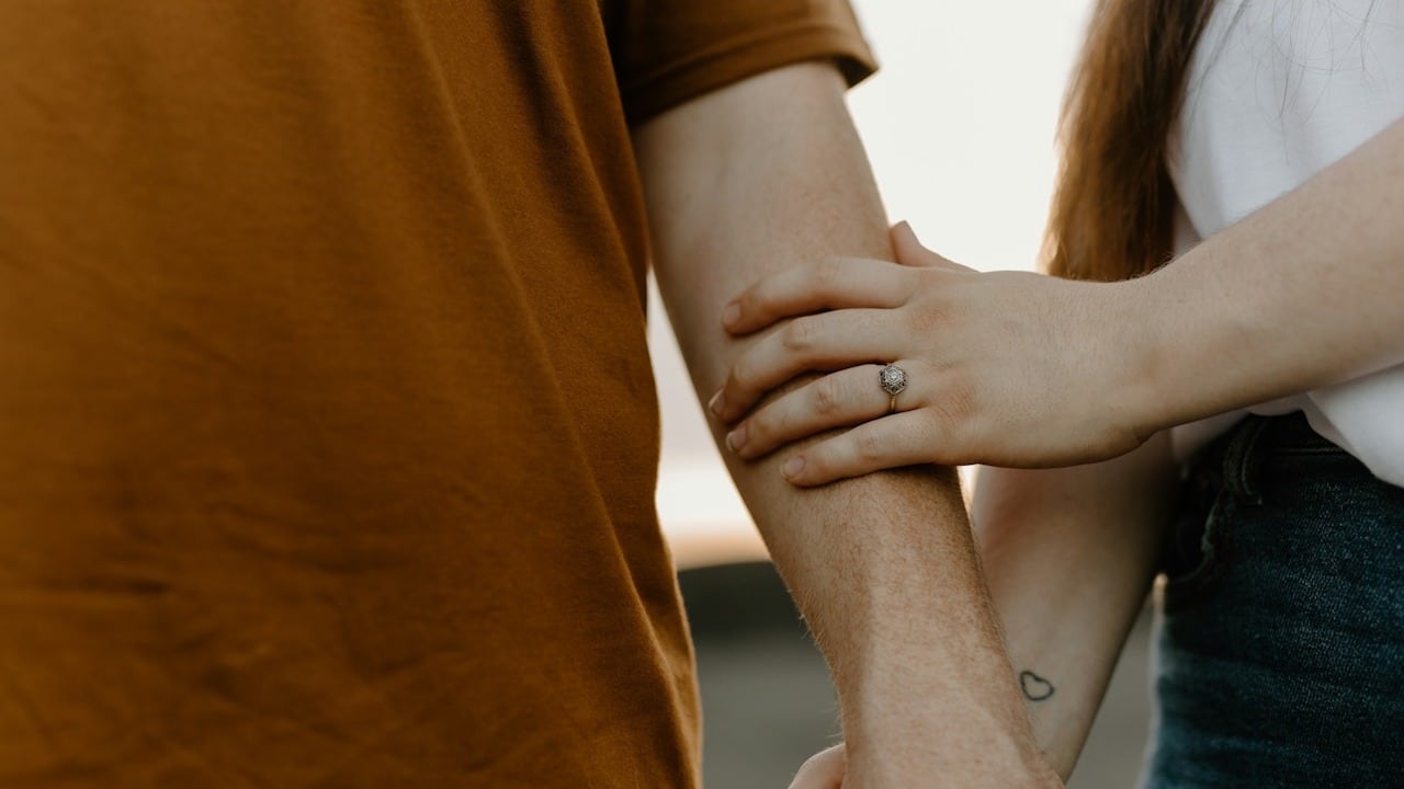 A close-up shot of a couple's hands; a woman's hand, featuring a ring and a small heart tattoo on the wrist, rests gently on a man's forearm as they hold hands.