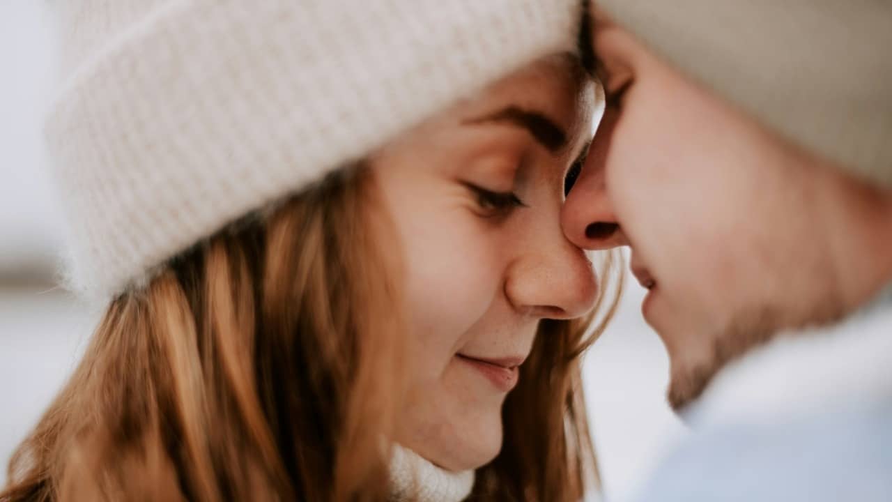 A close-up shot of a couple wearing beanies, leaning in with their foreheads and noses touching.