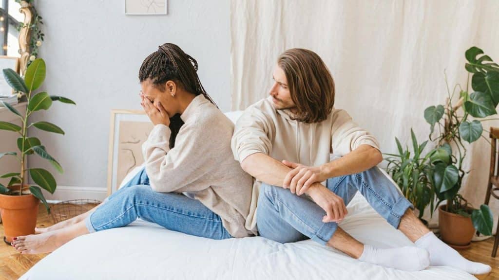 A woman sits on a bed covering her face while a man sits beside her looking away.