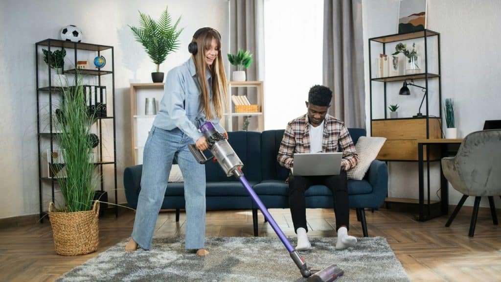 A woman vacuums a rug while a man sits on a sofa using a laptop computer.