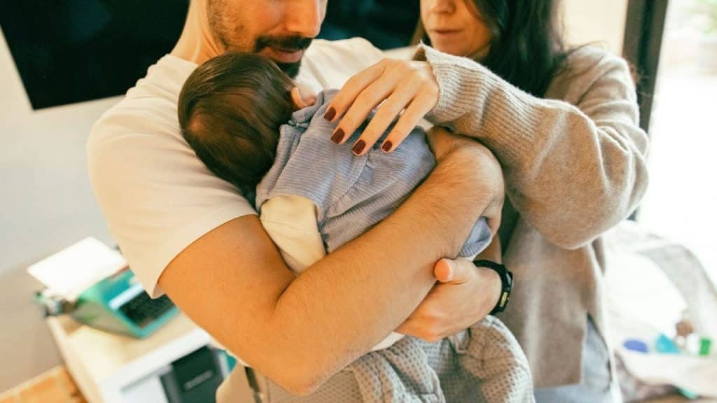 A bearded man holds a sleeping infant while a woman reaches out to touch the child.