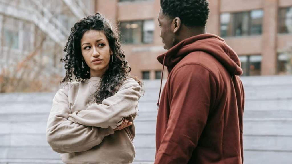 A woman with curly hair stands with crossed arms while looking sideways at a man.