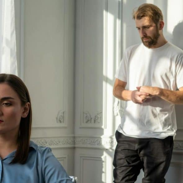 A woman in a blue shirt looks away while a bearded man stands against a wall.