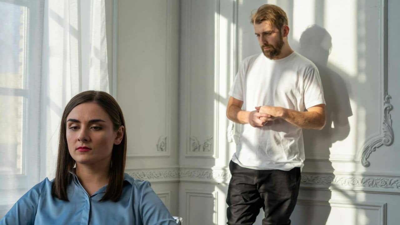A woman in a blue shirt looks away while a bearded man stands against a wall.