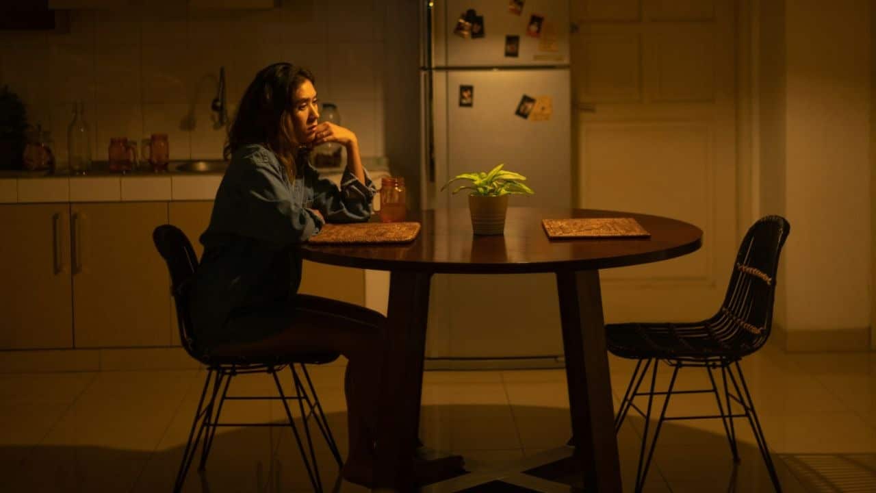 A woman sits alone at a wooden table in a dimly lit kitchen at night.