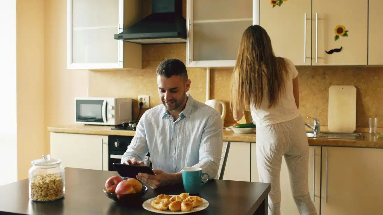 A man uses a tablet at a table while a woman works in the kitchen.