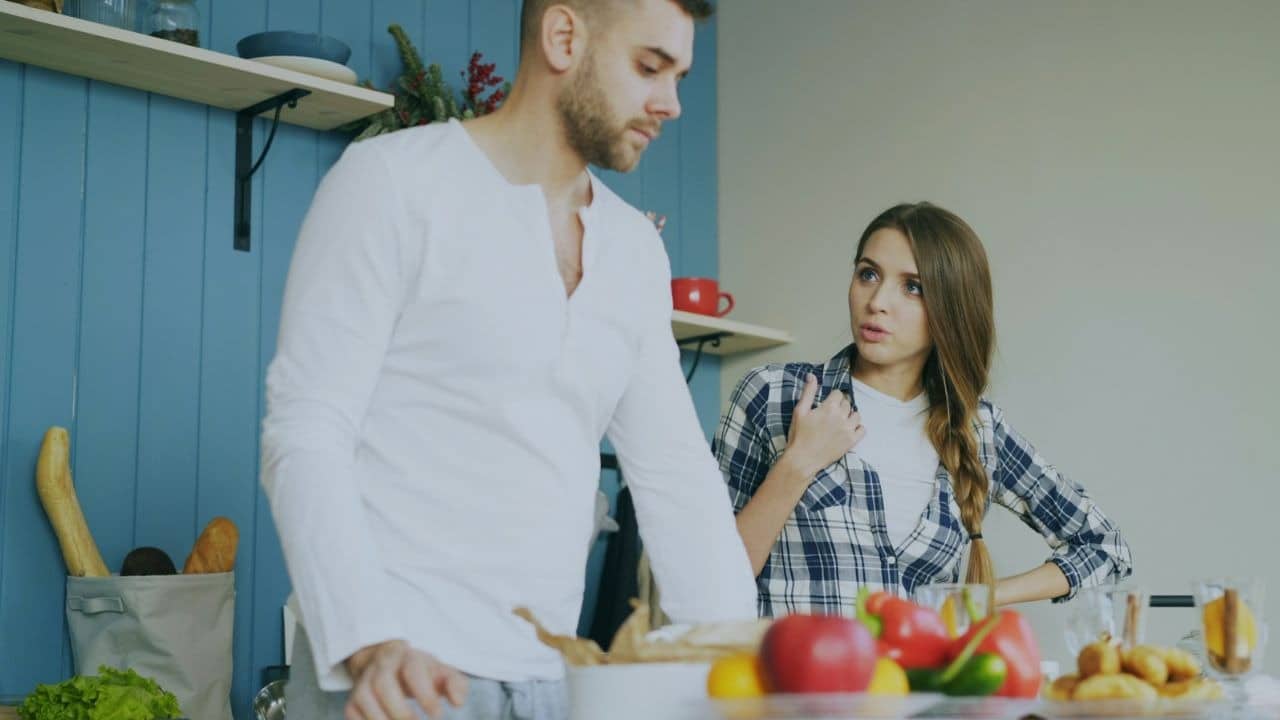 A woman in a plaid shirt speaks to a man standing in a kitchen area.