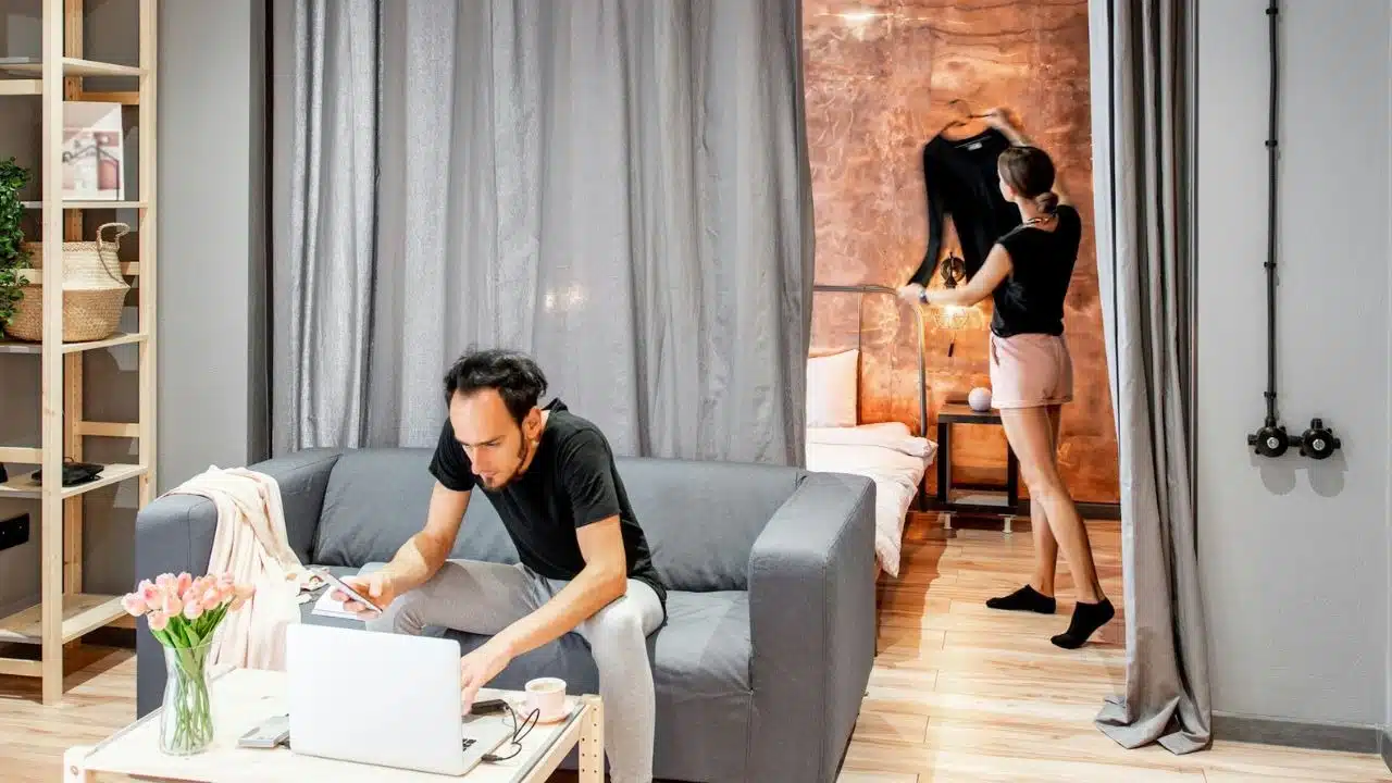 A man works on a laptop while a woman organizes clothes in the background.