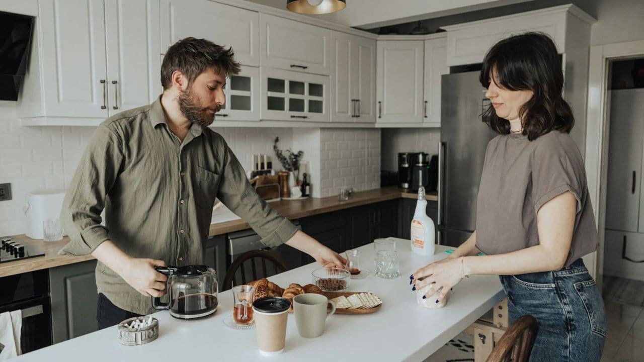 A man and woman stand in a white kitchen preparing food and pouring coffee.
