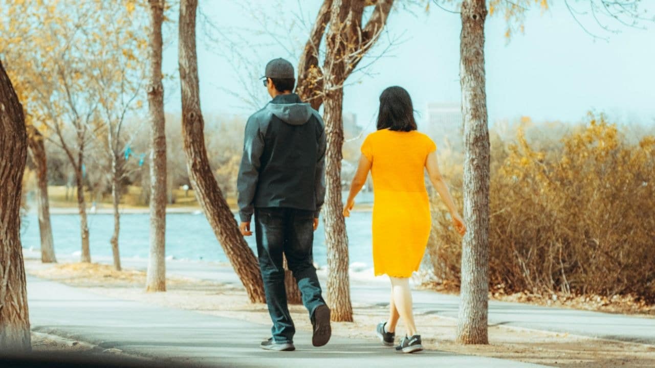 A man and a woman in a yellow dress walk away on a paved path.