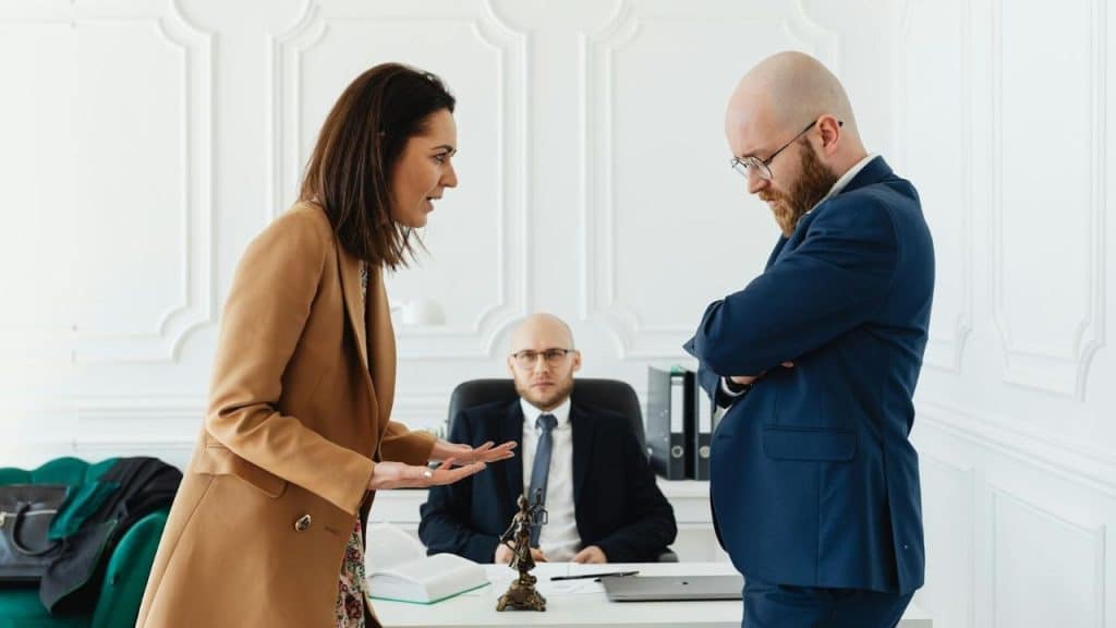 A woman gestures toward a man with folded arms while a seated man watches them.