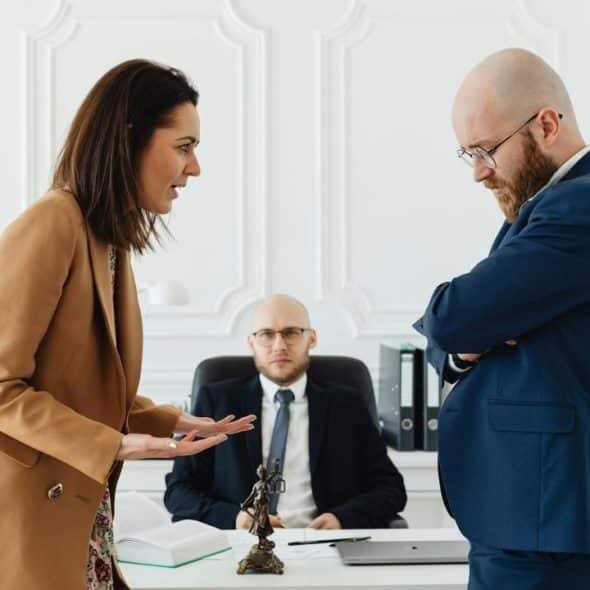 A woman gestures toward a man with folded arms while a seated man watches them.