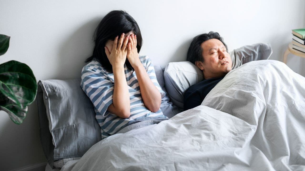 A woman covers her face while sitting in bed next to a man who is sleeping.