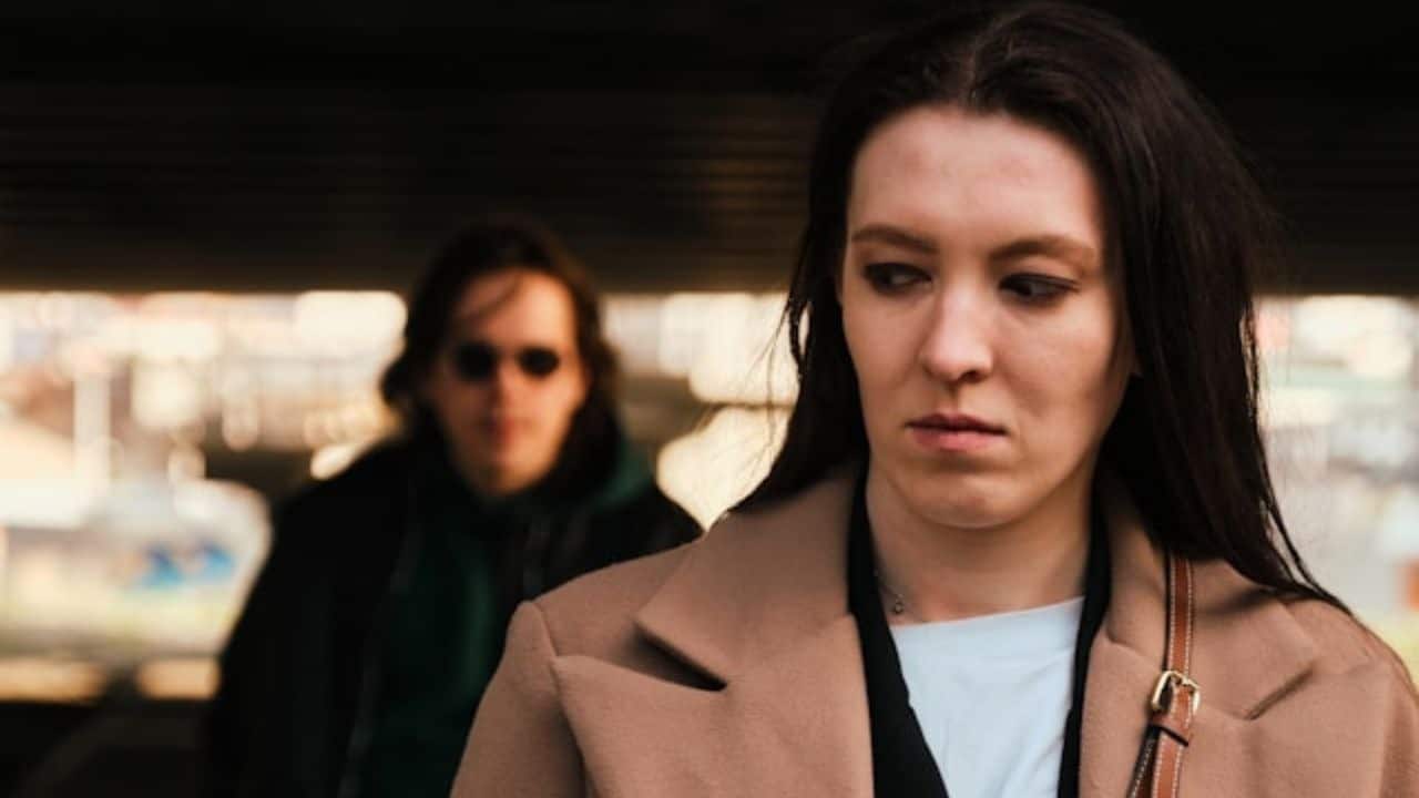 A woman in a tan coat looks down as a blurred man follows behind her outdoors.