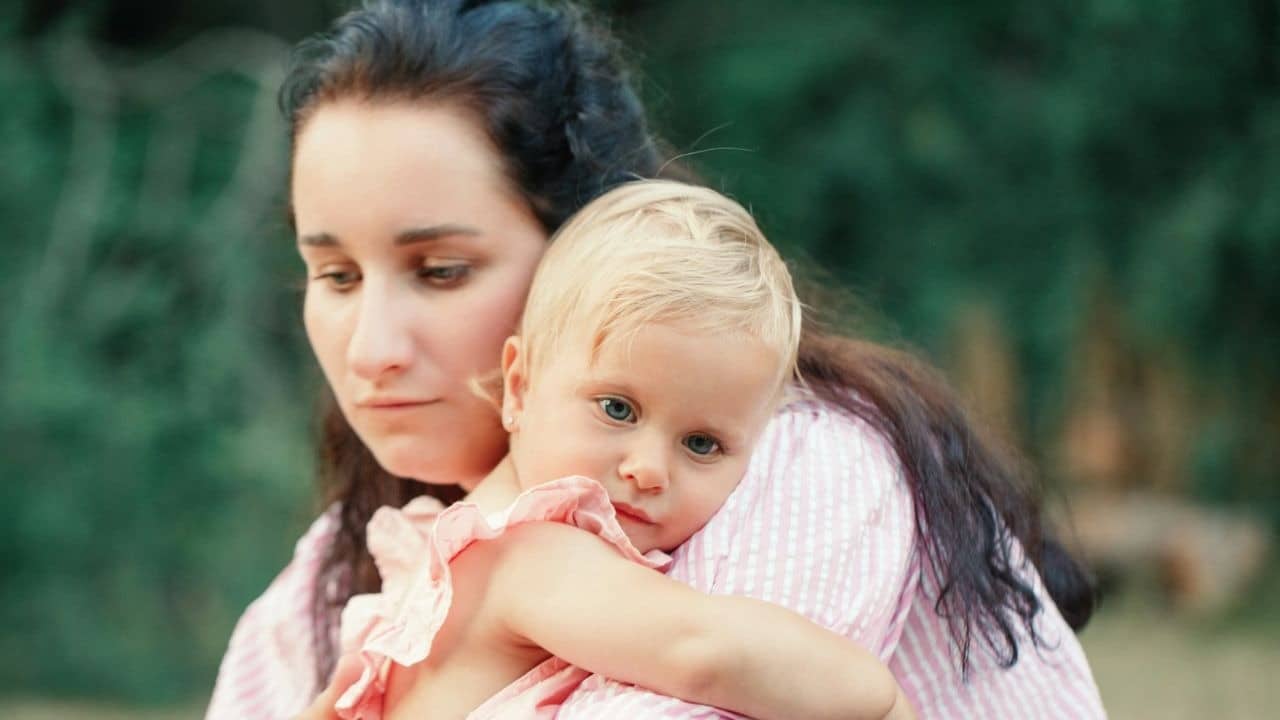 A woman with long dark hair holds a small child wearing a pink ruffled shirt.