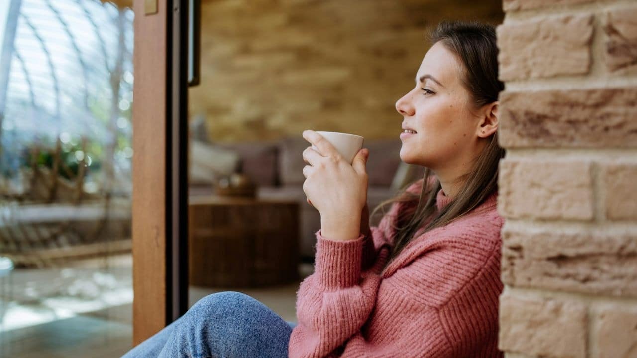 A woman in a pink sweater sits on the floor holding a white mug near a window.