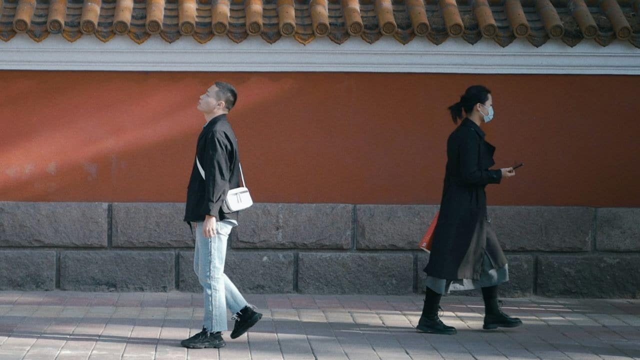 A man and a woman walk in opposite directions past a long orange and grey wall.
