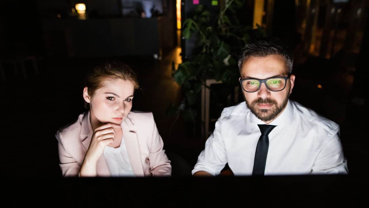 A man and woman look at a bright screen in a dark room with green plants.