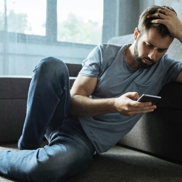 A man with a beard sits on a carpeted floor, leaning against a gray couch.