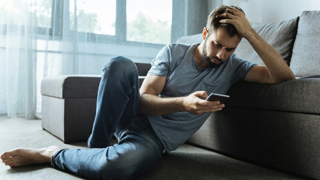 A man with a beard sits on a carpeted floor, leaning against a gray couch.