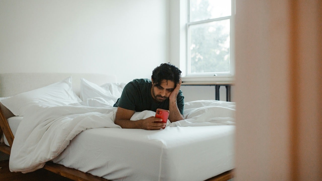 A man lies on a bed with white linens, holding a red smartphone in a room.