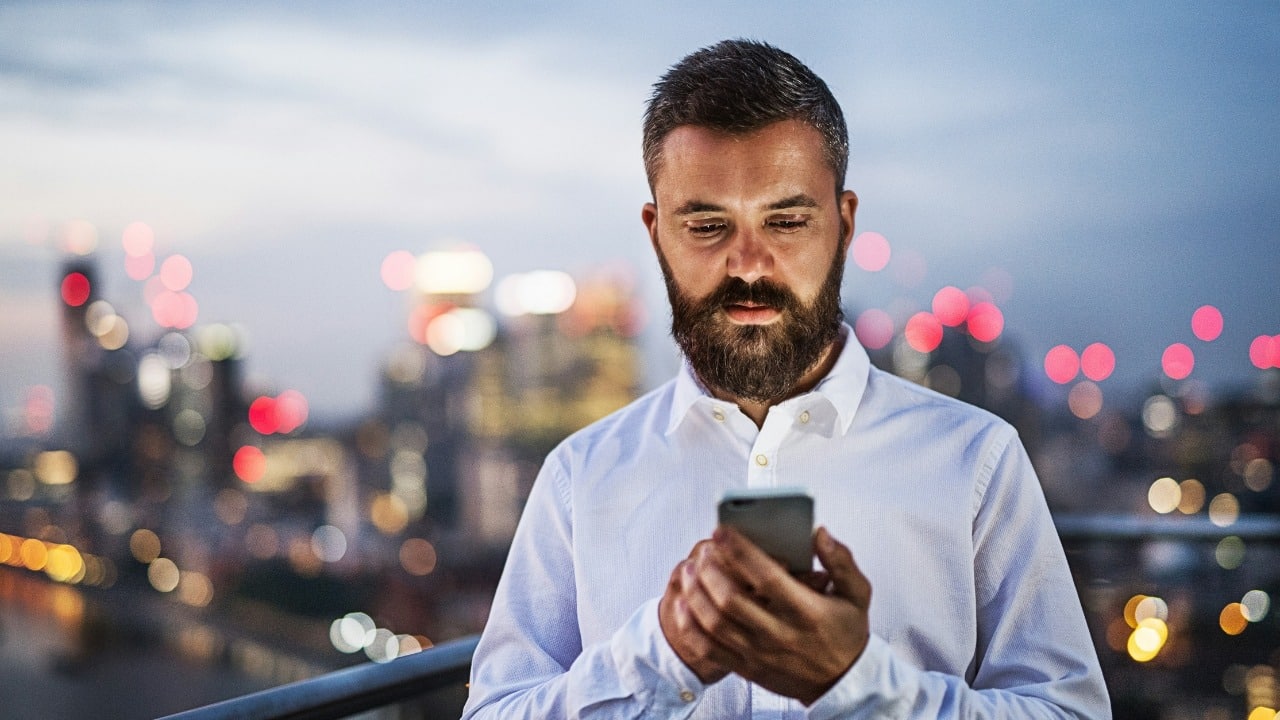 A bearded man in a white shirt looks down at a phone with city lights behind.