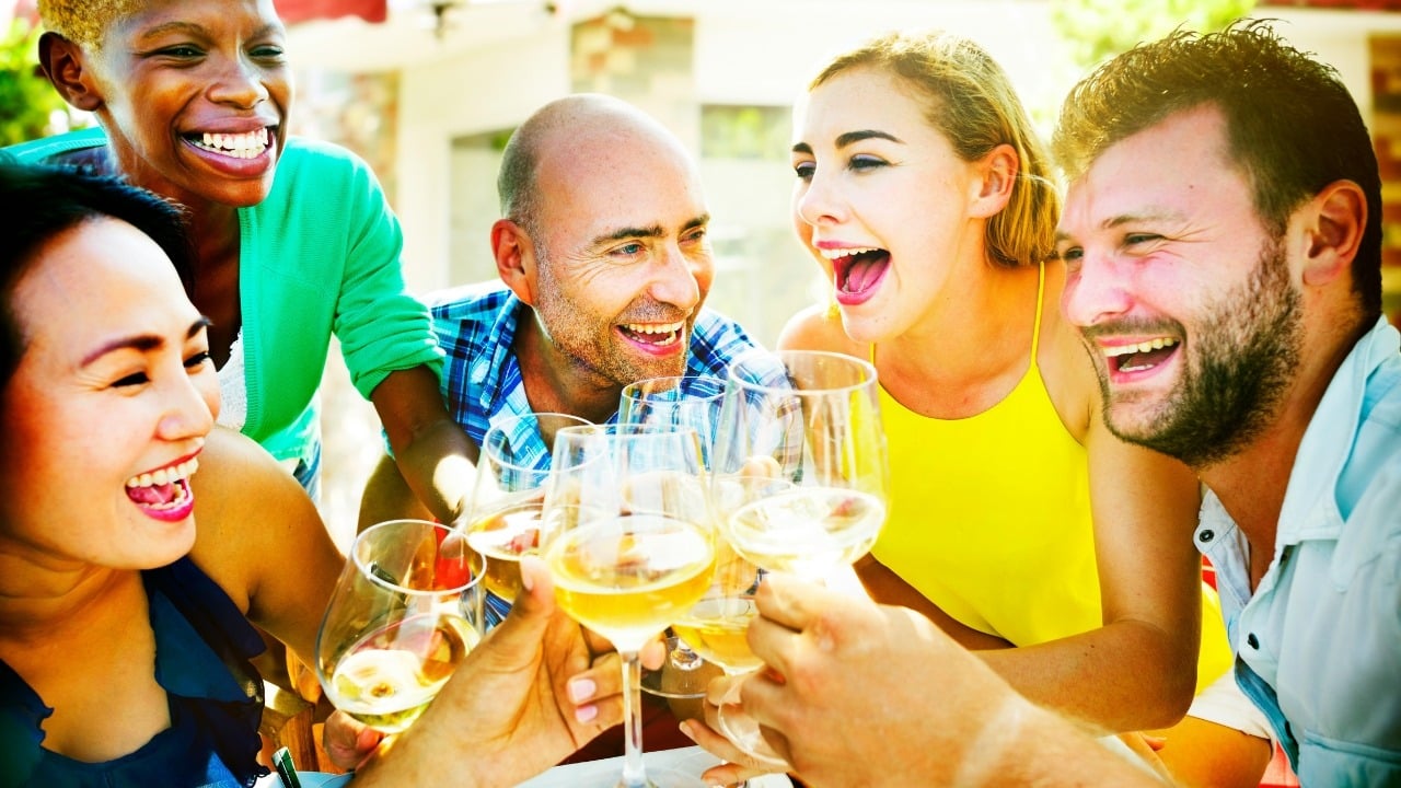 A group of diverse people smile and cheer while clinking wine glasses together outdoors.
