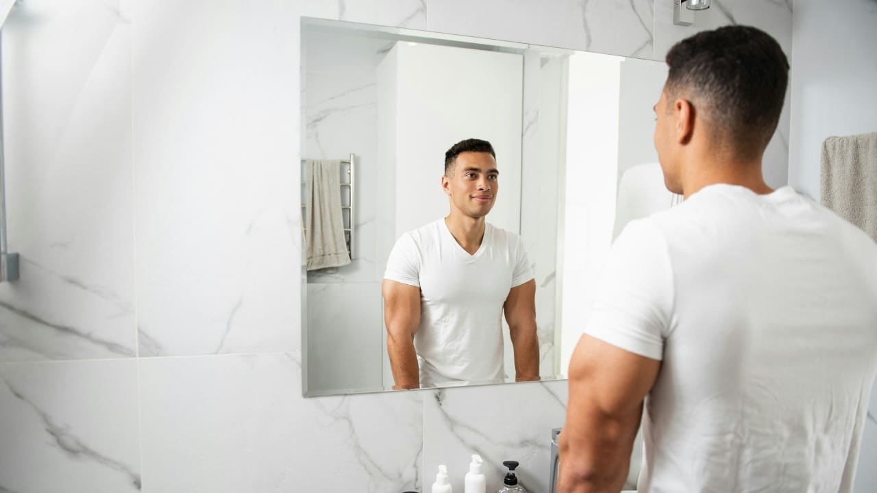 A man in a white t-shirt looks at his reflection in a large bathroom vanity mirror.