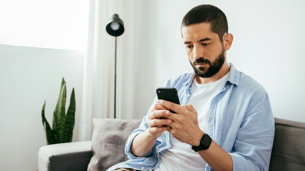 A bearded man in a blue button-down shirt sits on a couch looking at a phone.