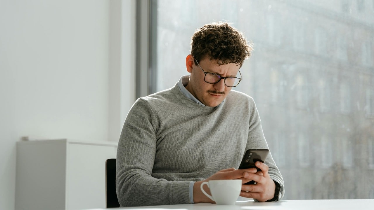 A man with glasses and a mustache looks at a smartphone while sitting at a table.