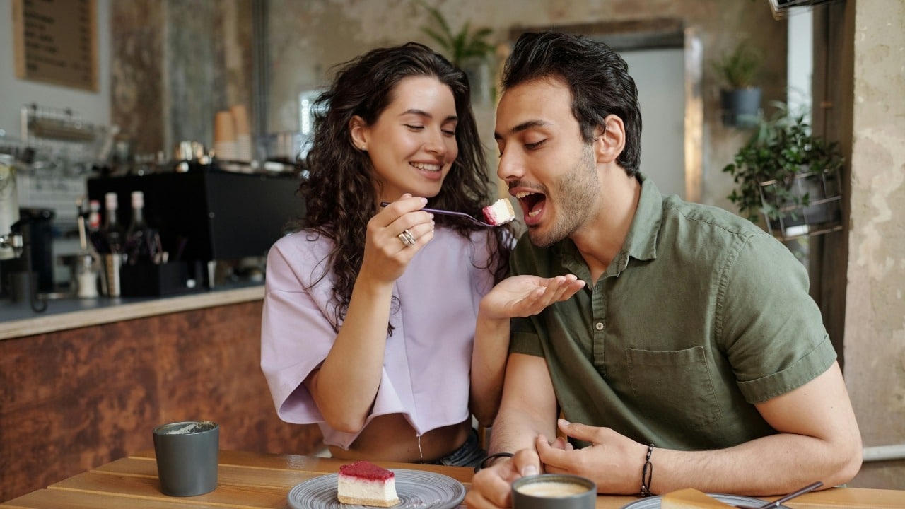 A woman feeds a piece of cake to a man sitting next to her in a cafe.