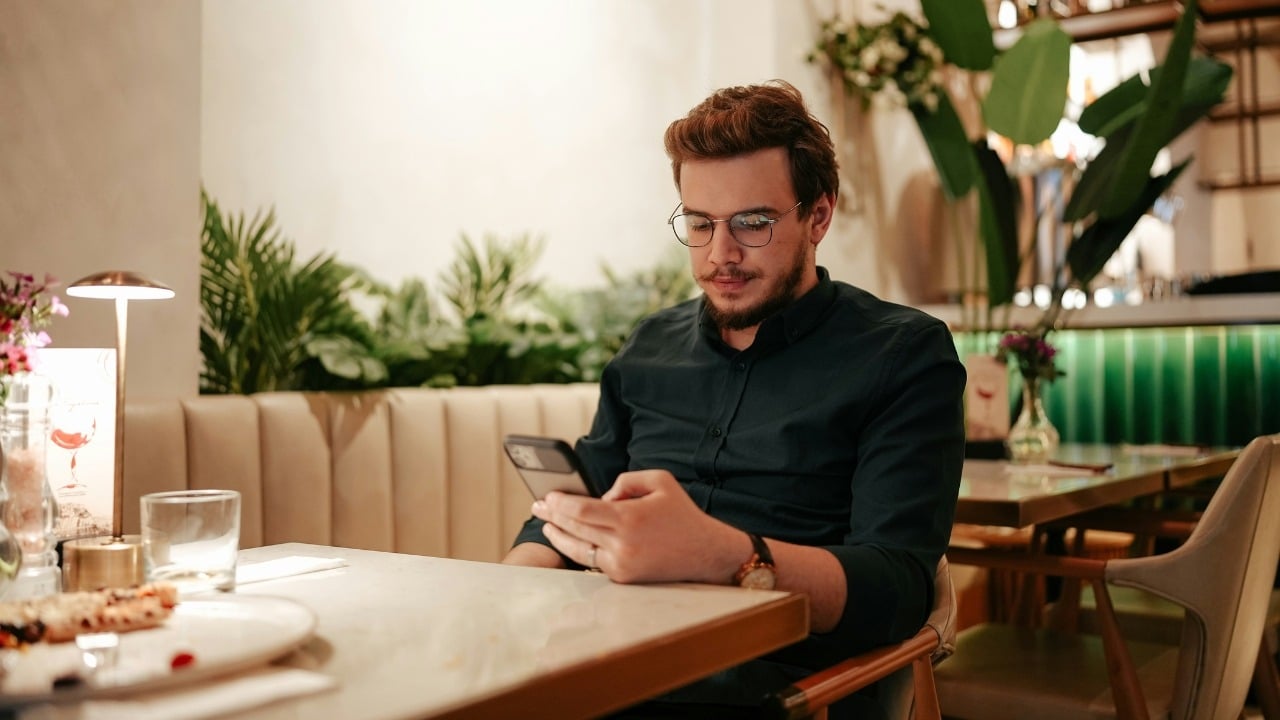A man with glasses and a dark shirt sits at a restaurant table using a phone.