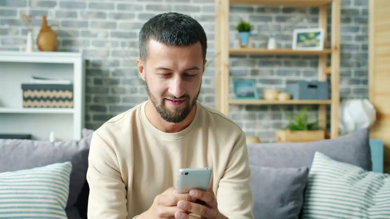 A bearded man in a tan sweater sits on a couch and looks at a smartphone.