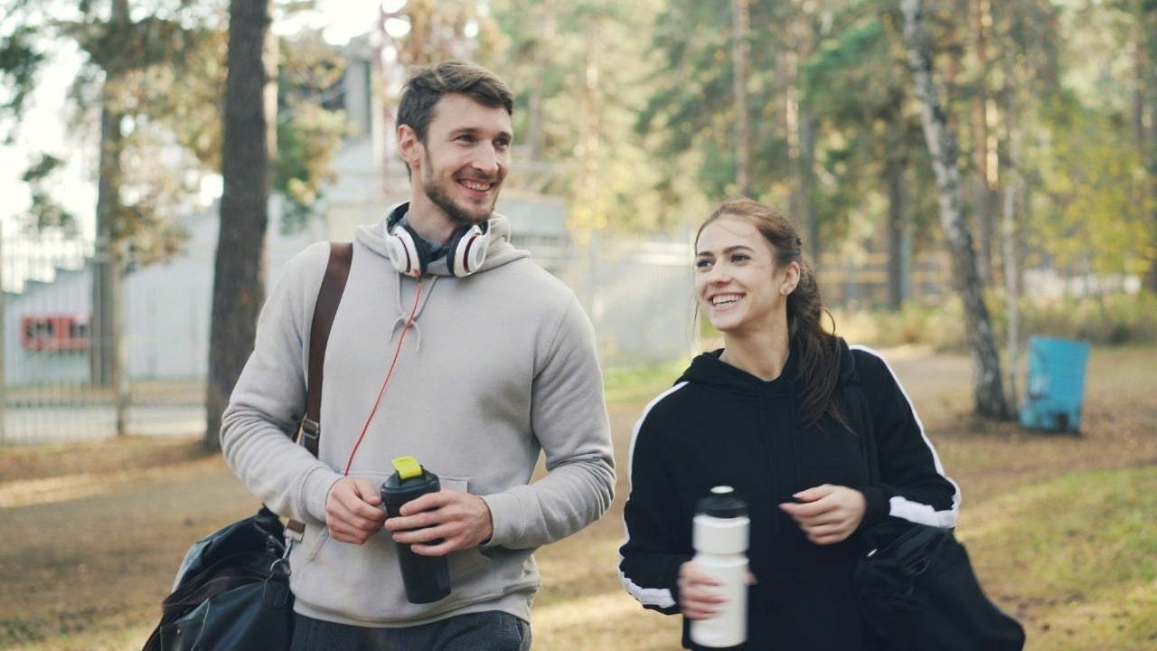 A man and woman in athletic wear walk through a park while smiling and holding bottles.