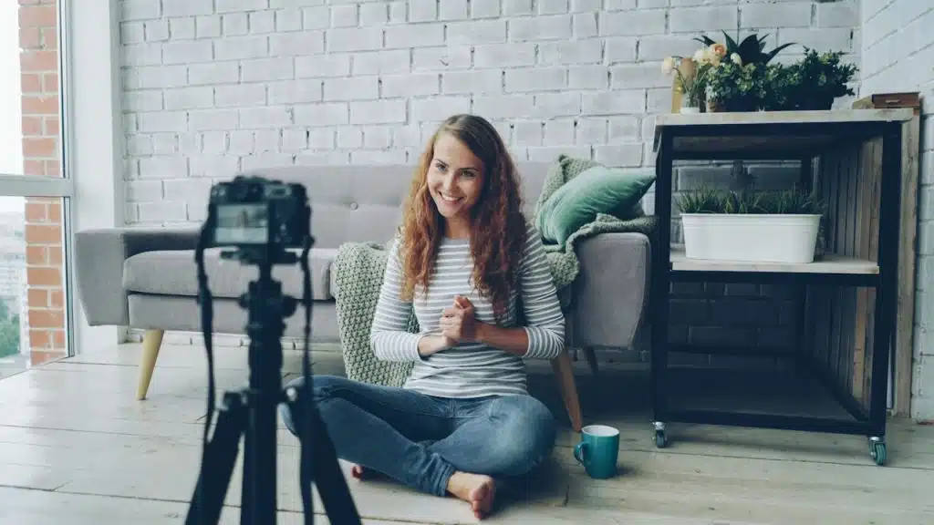 A smiling woman with long curly hair sits cross-legged on the floor of a bright living room, facing a camera on a tripod as if recording a video.