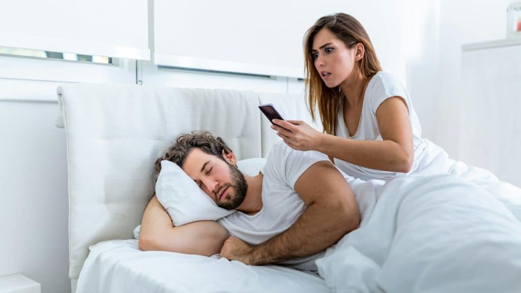 A shocked, jealous wife spies on the phone of her husband while the man is sleeping in bed at home.