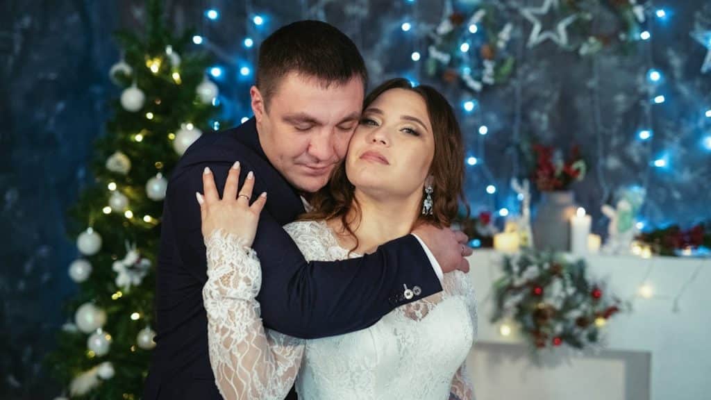 The man hugs the woman from behind with his eyes closed, while she looks calmly toward the camera.