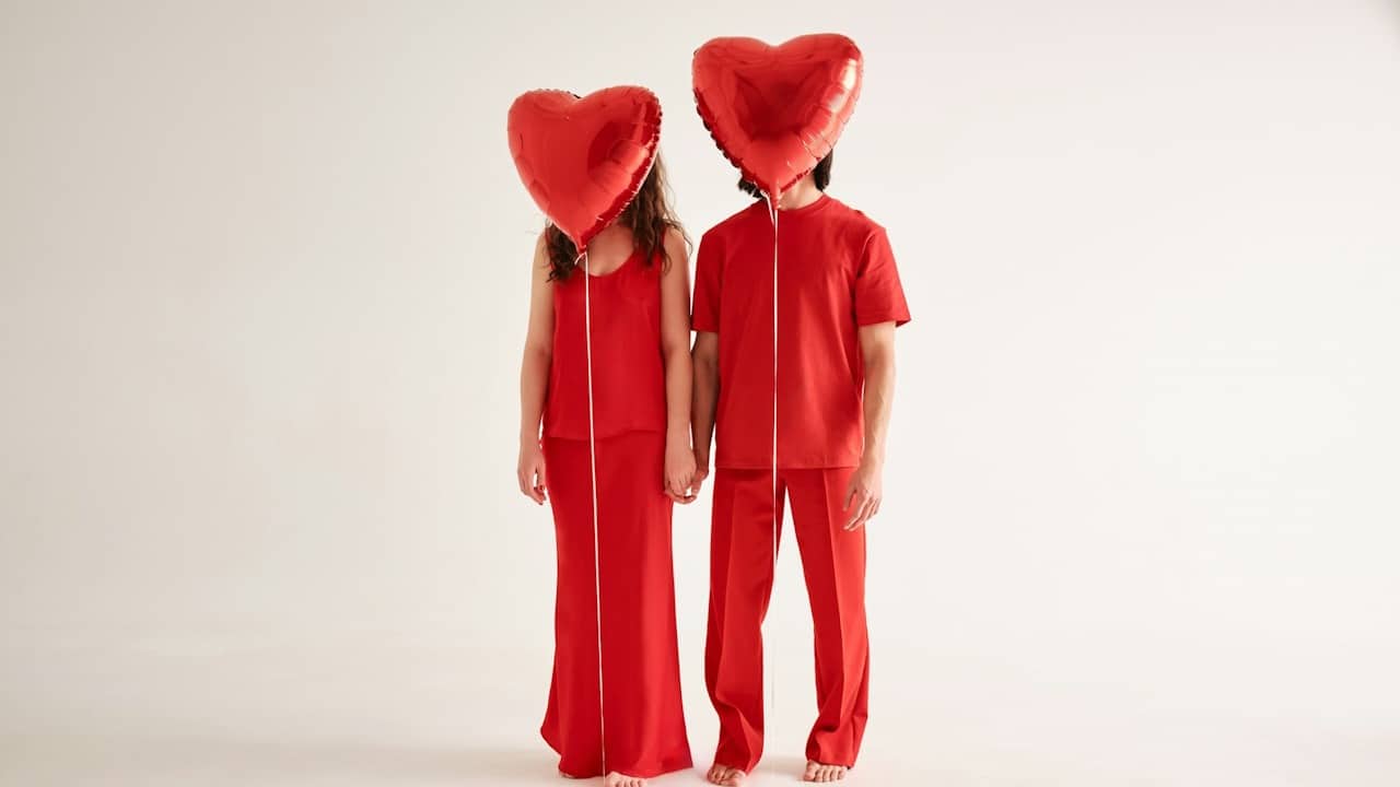 A couple with red dresses with faces hiding with red heart balloons.
